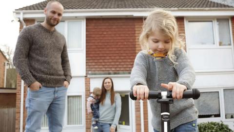 A mother and father outside a brick house with a small child and a baby