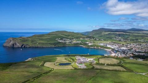 An aerial view of Port Erin showing a blue bay with a beach surrounded by rolling green hills and green fields. There is also a hub of white houses to the right of the beach and a large development on the headland in the fore.