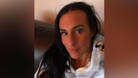 A selfie of Lisa Denton - she has long dark hair and stares into the camera. She is pictured sitting on a sofa, wearing a fluffy hoodie decorated with dogs. 
