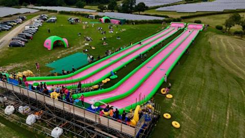 The giant green and pink water slide in a large field. There is cars parked in the car park next to it.