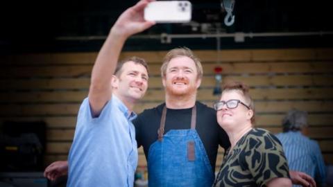 Jack Stein smiles as a man and a woman take a selfie with him. He has fair hair and a ginger beard, and is wearing a black T-shirt and blue apron. The man taking the selfie is holding the phone in his right hand.