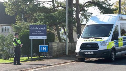 A female police officer stands at the entrance to Gainsborough Care 鶹Լ. A marked police van is parked on the roadside. The two-storey building can just be seen behind some trees and a blue sign saying 'Gainsborough Care 鶹Լ'.