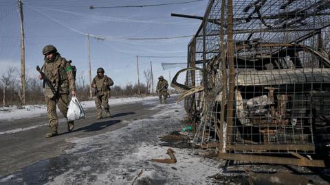 Three soldiers walk on an icy road in their military fatigues carrying weapons and sacks. A burnt out car in a cage is on the side of the road on the right.