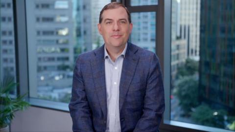 A medium close up of Matt Garman, chief executive Amazon Web Services, sat facing the camera in front of a window of an office building with the city outside visible. He is smiling slightly and wearing a blue suit and a shirt without a tie.