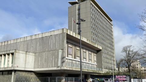 A grey tower block with a lower grey 1960s building to its left is photographed against a pale blue sky. A Union Flag is flying form a flagpole on the lower building.