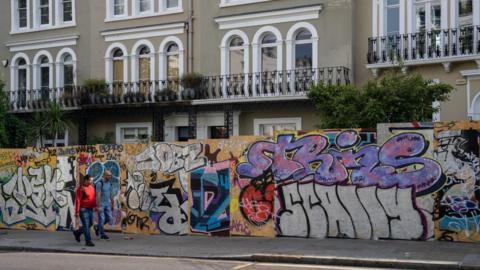 Colourful chipboard panels covering the length of pavement and behind there are terraced houses. Two men wearing jeans and casual tops, one red long sleeved top, the other with a blue t-shirt, are walking by.