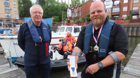 Two men, wearing life jackets, are standing on a dock. The one on the left, balding with a red beard, is smiling and holding a case containing a medal. There is another medal on a ribbon around his neck. The other man has short grey hair and is wearing spectacles. Behind them there are two men on a small boat, and a larger craft behind them.