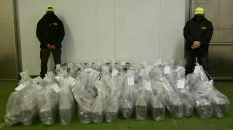 Two officers wearing black jackets and trousers and black face masks and luminous yellow police baseball caps to disguise their identity stand behind dozens of plastic sacks containing seized drugs.