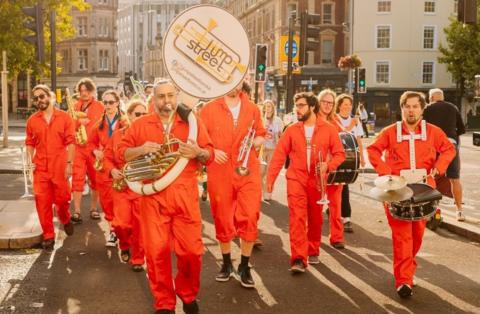 Musicians in orange jump suits parade in Bristol city centre ahead of the Proms coming to the Bristol Beacon. They are holding different instruments including saxaphones and trombones