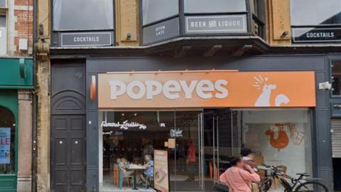 The shop front of Popeyes Louisiana Chicken restaurant on Oxford's Queen Street. Some people are outside in front of it while others are eating inside.