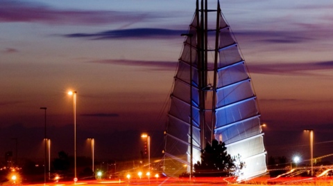 The Tri-Sail sculpture, which has sails like a yacht, pictured during an orange sunset and also showing the lamposts and other lights in the area lit up, creating an orange glow