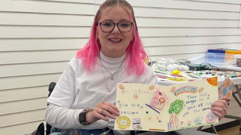 Katie Callaghan is wearing a white roll neck and has pink hair. She is sat at a black table which is covered in handmade cards. She is holding one open towards the camera.