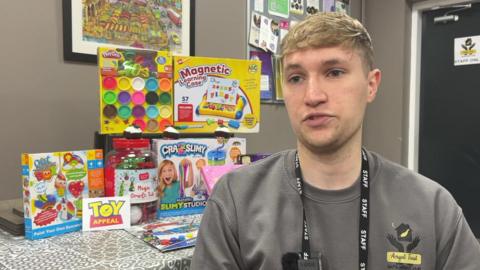 Elliot Pole sitting in front of a table which has toys and games stacked up. Mr Pole has short fair hair and is wearing a grey jumper with the Angel Trust logo. 