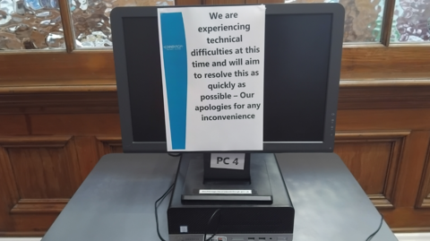 A black desktop PC screen at a library in Edinburgh which has been turned off and has an A4 notice taped to it. The notice reads: "We are experiencing technical difficulties at this time and will aim to resolve this as quickly as possible - Our apologies for any inconvenience". The screen is on a grey table in front of a wooden background with frosted glass windows.