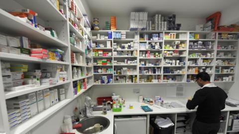 A chemist works on prescriptions inside a pharmacy with shelves surrounding him full of medication.