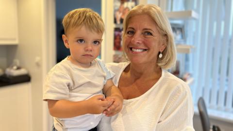 Photo of a woman with short blonde hair smiling at the camera. She is wearing a white T-shirt with silver earrings. She is holding a young boy with short blonde hair wearing a white T-shirt.