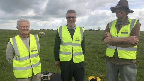 Three archaeologists standing in a field wearing hi-vis jackets