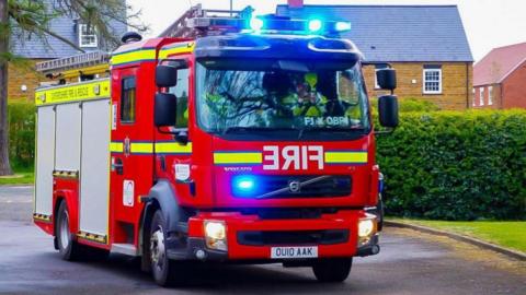 A red fire engine with blue emergency lights illuminated is driving along a residential street lined with brick houses and trees.
