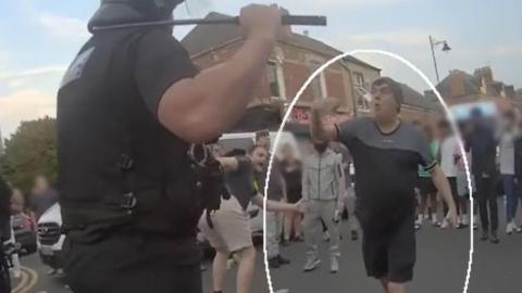 A still from police footage showing two men gesticulating at a police officer with a crowd behind them.