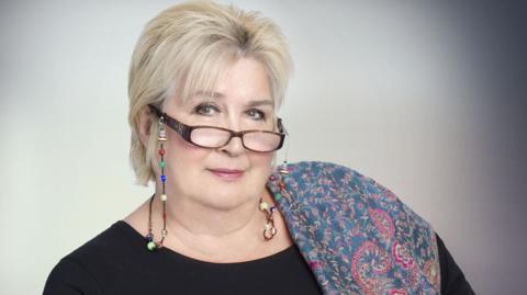 Dame Jenni Murray has a blue and red paisley pattened scarf over one shoulder and is wearing a black top, she has short hair and glasses which are perched on her nose and attached by a beaded string