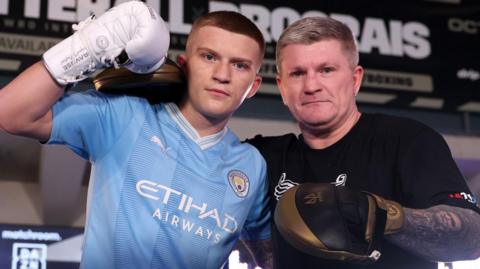 Campbell Hatton, wearing a blue Manchester City shirt and a white boxing glove, stands with his arm round his father Ricky Hatton, who wears a black T-shirt and a black and gold sparring glove