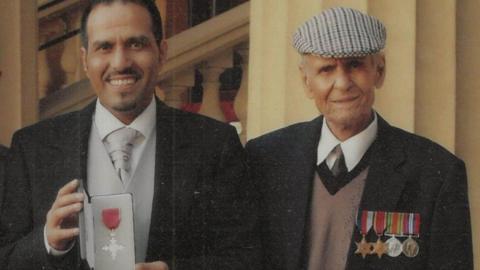 Mashuq Hussain holding his OBE in grey and black tie and tails, standing next to  Ghulam Hussain wearing his medals  