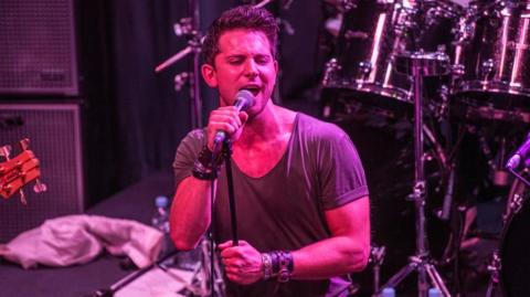 Man with brown hair sings into microphone on stage with red lighting shining down on him. He wears a tan t-shirt and black punk wristband.
