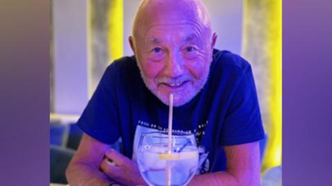 A man wearing a navy blue T-shirt is smiling at the camera while leaning on a table. In front of him is a cocktail with a straw. There is ambient yellow lighting behind him.