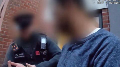 A screenshot from a police body camera of a man being handcuffed by a police officer in a grey uniform. Both the man and the police officer's faces have been blurred out. 