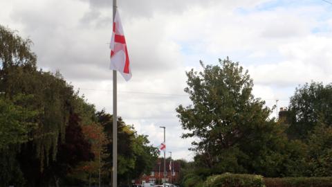 St George cross flags hung on a row of lamp-posts with trees on either side of the road.