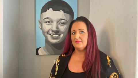 Dionne Barrett, who has long dyed purple hair, stands in front of a print of her son, Gordon Gault, on the wall. He is coloured grey and on a blue background. He is smiling.