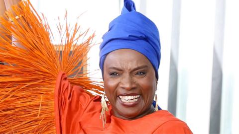 Angélique Kidjo smiles and lifts one arm in a long-sleeved dress with tasselled sleeves and a royal blue headwrap
