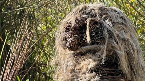 A figure made out of willow and hemp, shaped to create eyes, a nose and straw-like hair, against a backdrop of reads and foliage.