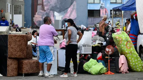 People gather with their belongings near a mural of late Venezuelan President Hugo Chavez,