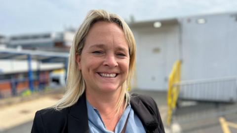 Ms Grimmett with long blond hair, wearing a dark blue jacket and blue shirt with a building site behind her