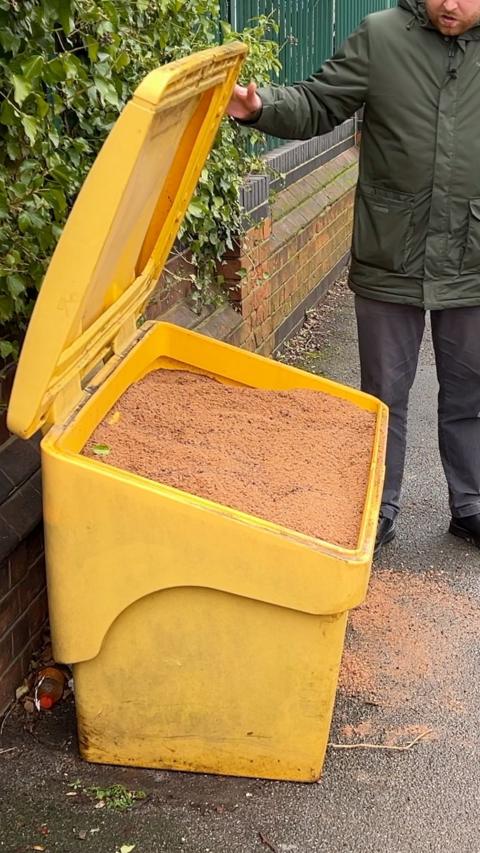 A large yellow roadside grit bin with its lid open. The bin is filled to the top with reddish-brown grit and some of the material has spilled onto the pavement in front of it. The bin is positioned next to a brick wall and green foliage. A person is standing to the right side of the bin, wearing a dark green coat and dark trousers. Only the lower part of the person’s face and body is visible. The person is holding the grit bin lid open with one hand.