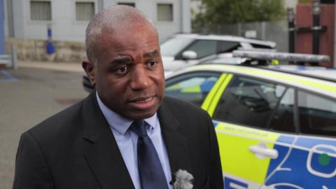 Close up of David Lammy in black suit, shirt and blue tie as he speaks to the press while standing in front of a police car in the street