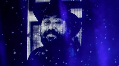 Picture of Gearóid Ó Cairealláin that was displayed on a screen in the SSE Arena during a gig by Kneecap - he has a dark beard and is wearing a wide-brimmed had