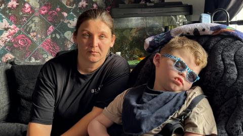 There is a woman with tied back black hair with a black tshirt on. She is sat on a grey sofa. To her left, there is a boy with brown hair, blue and white rimmed glasses and a light brown t-shirt. He is sat down on a black comfortable chair, and his head is tilting to the left.