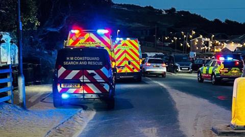 Several emergency vehicles on a beach road a night - with their lights on.