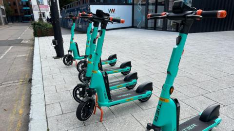 Seven e-teal-blue coloured e-scooters lined up in a row on a city centre pavement.