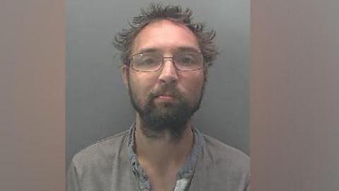 Police mugshot of a man with grey sticky-up hair and glasses. He is wearing a grey open-necked shirt