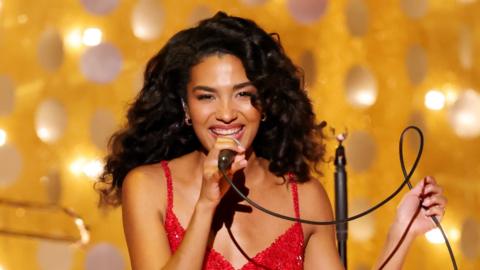 Olivia Dean holds a microphone and smiles as sings on stage. She's wearing a red, sequin dress and is stood in front of a golden background.