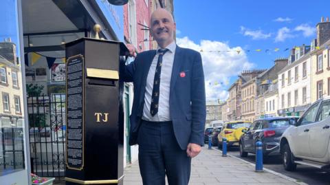 A bald-headed man wearing a suit leans against a black post box, with a row of cars behind him