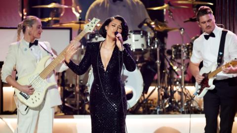 A front on photo of a singer and two guitarists on a stage. The singer is in the middle wearing a black, long sparkly dress. She has short black hair and is in the middle of singing a song. Either side of her are two guitarists. On the left the woman is wearing a white suit holding a white guitar and the man on the right is wearing black trousers and a white shirt.