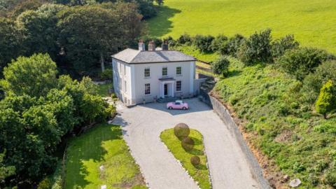 Drone shot of Penally House. It is a two-storey country house set in a green landscape. The house is light-coloured, with symmetrical windows and a classical entrance featuring columns and a porch. A winding gravel driveway leads up to the front of the house, where a small pink car is parked. The property is surrounded by manicured lawns and trees, with rolling hills in the background. In the centre of the driveway, there are three spherical metal sculptures arranged in a line.