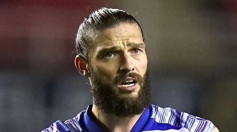 Andy Carroll during a game. He is wearing a blue and white-striped football kit. He has his black hair pulled back into a bun and has a bushy dark beard. He is sweaty during a football match.