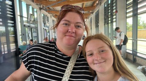A close up shot of a mother standing with her her daughter in a modern, glass-fronted bus station. The mother has reddish-brown hair, tied back, with sunglasses on the top of her head and is wearing a black T-shirt with white stripes. Her daughter has long strawberry blond hair, is wearing a white top and is smiling.