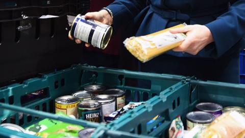 Two plastic crates have tins and other food packets in them.  Two hands are holding a tin and a packet of spagetti above the crates.