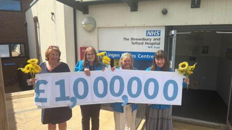 Four women are standing in a line and smiling. They are holding bunches of yellow flowers and a line of blue numbers on paper. The numbers read "one million pounds"